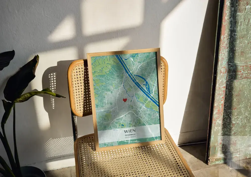 Grün-blaues Stadtposter mit Herz und hellem Koordinatenbanner im Naturholzrahmen auf einem Geflechtstuhl im Sonnenlicht